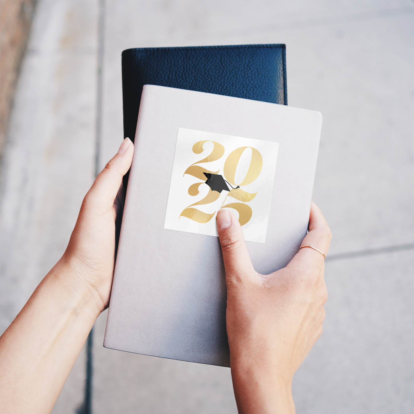 White square sticker with 2025 in gold and a black graduation cap placed in the middle stuck on a gray notebook being held.