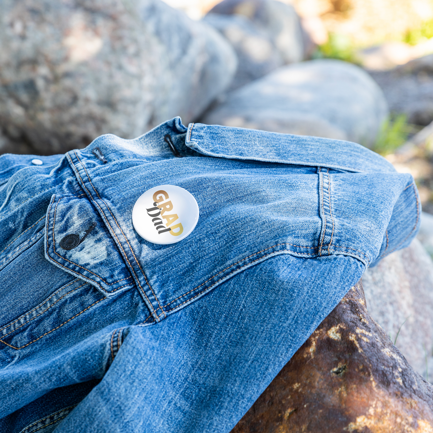 White button pin with Grad in gold and dad in black, pinned to a denim jacket laying on a rock.