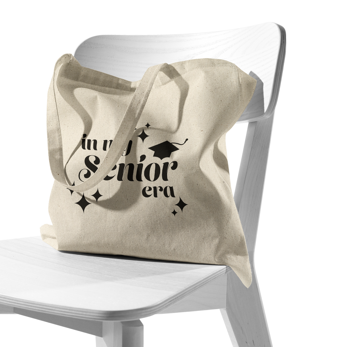 Tan canvas tote bag with the phrase “In My Senior Era” and a graduation cap printed in black ink sitting in a white chair.