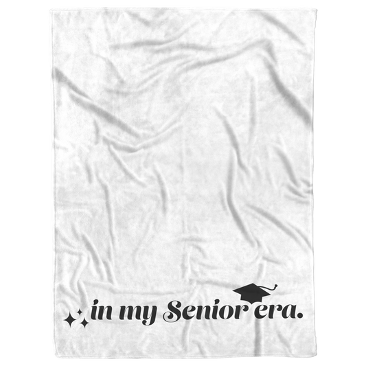 A white fleece blanket with the phrase "In My Senior Era" and a graduation cap in black at the bottom laying flat.