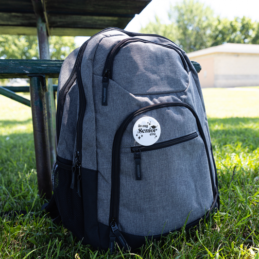 White button pin with the phrase "In My Senior Era" and a graduation cap in black pinned to a gray backpack sitting in the grass next to a picnic table.