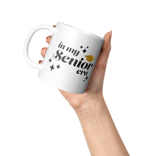 11oz white mug with the phrase “In My Senior Era” in black ink and a gold graduation cap being held by one hand at an angle.
