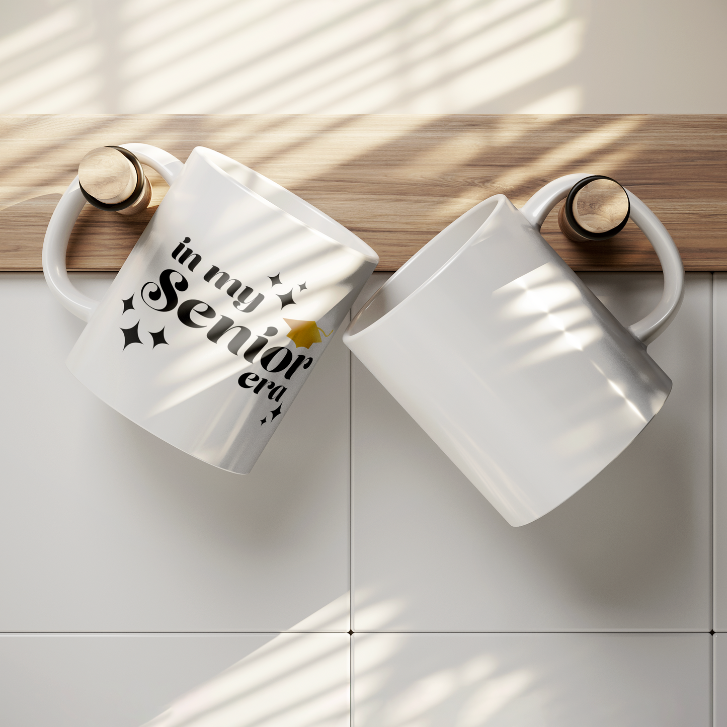 11oz white mug with the phrase “In My Senior Era” in black ink and a gold graduation cap hanging from a hook on a wall next to a blank white mug.