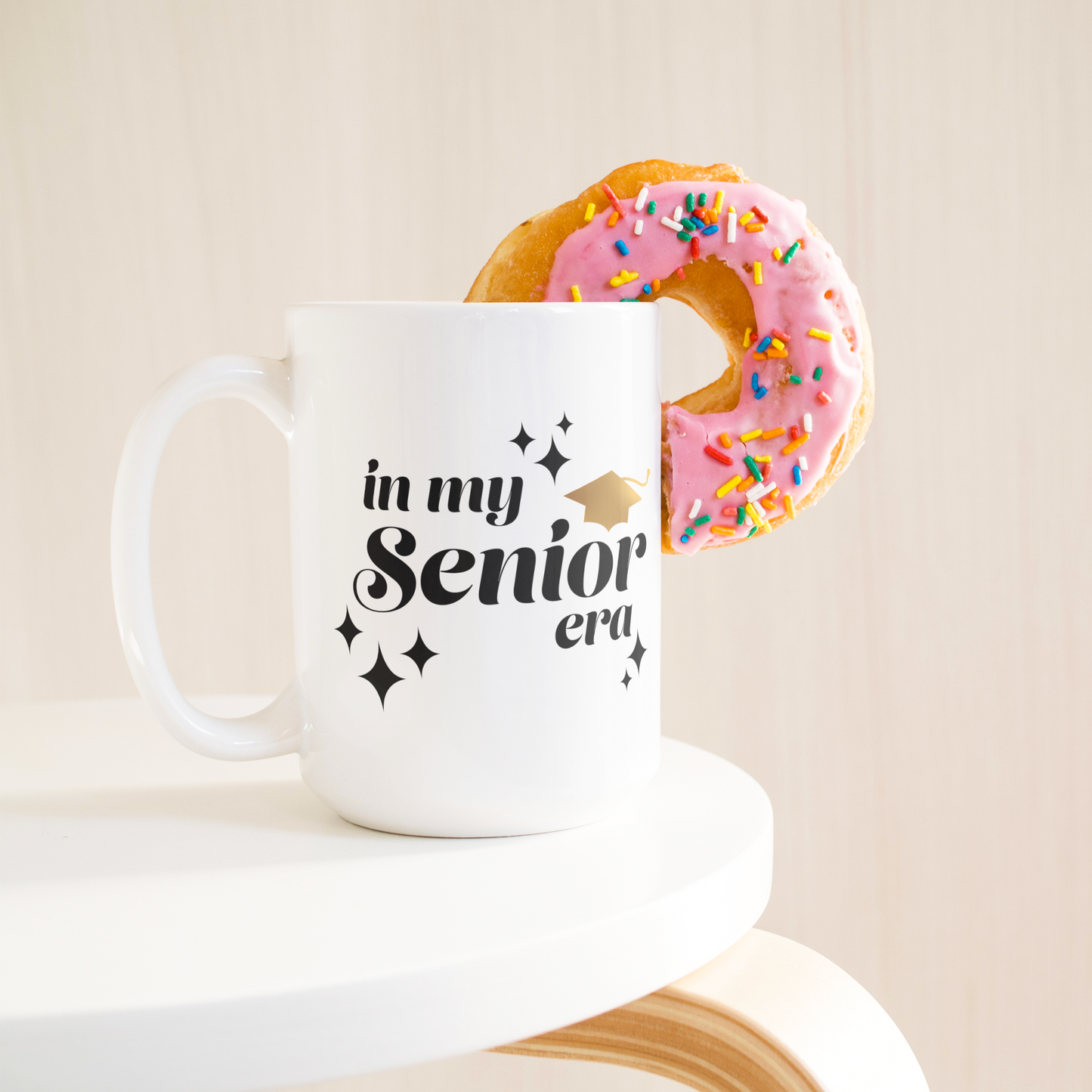 15oz white mug with the text "In My Senior Era" in black and a gold graduation cap sitting on a table with a donut sticking out of the mug.