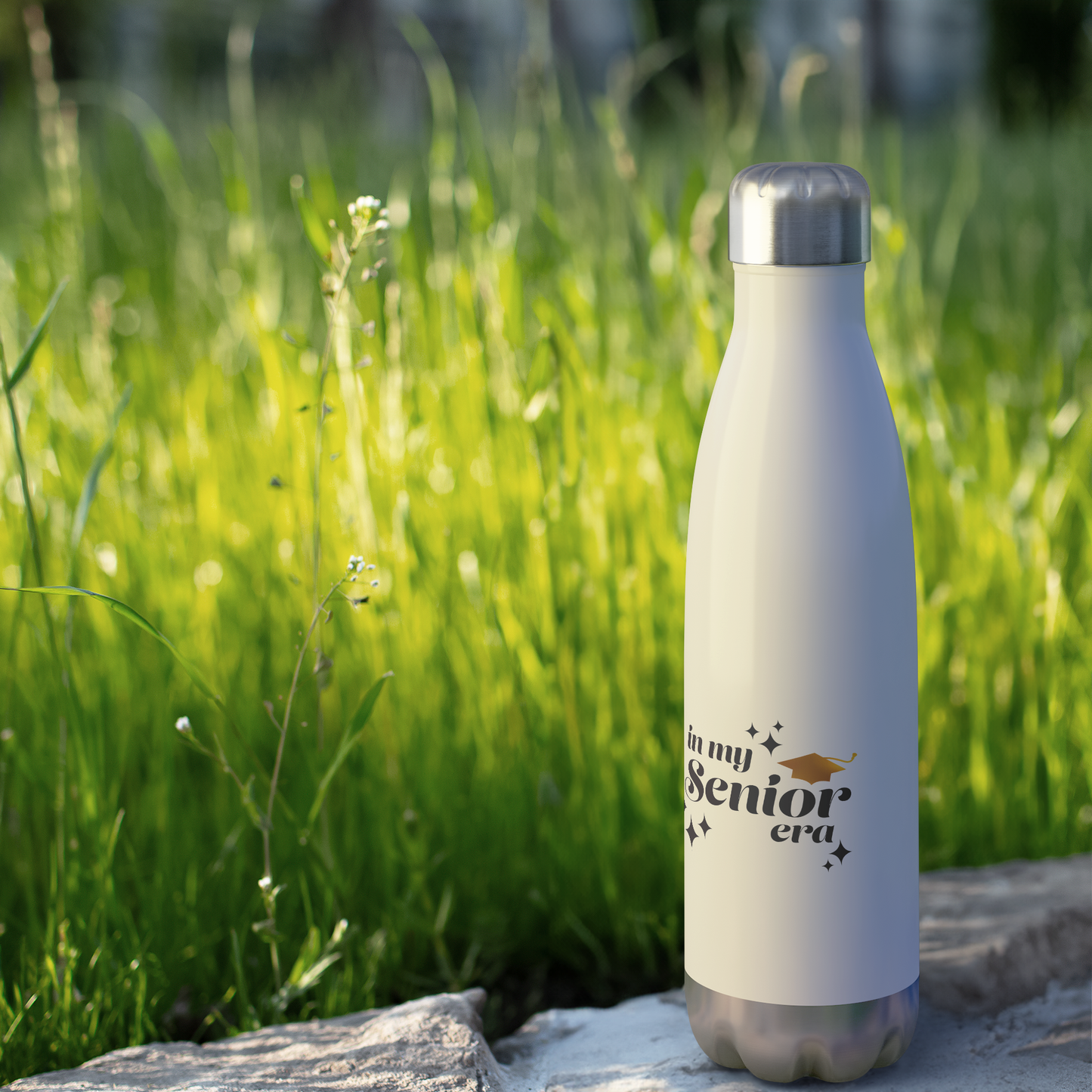 A white, 20oz water bottle with a silver lid and base sitting on a rock by grass. Text in the bottom center of the bottle says "In my Senior Era" in black with a gold graduation cap.