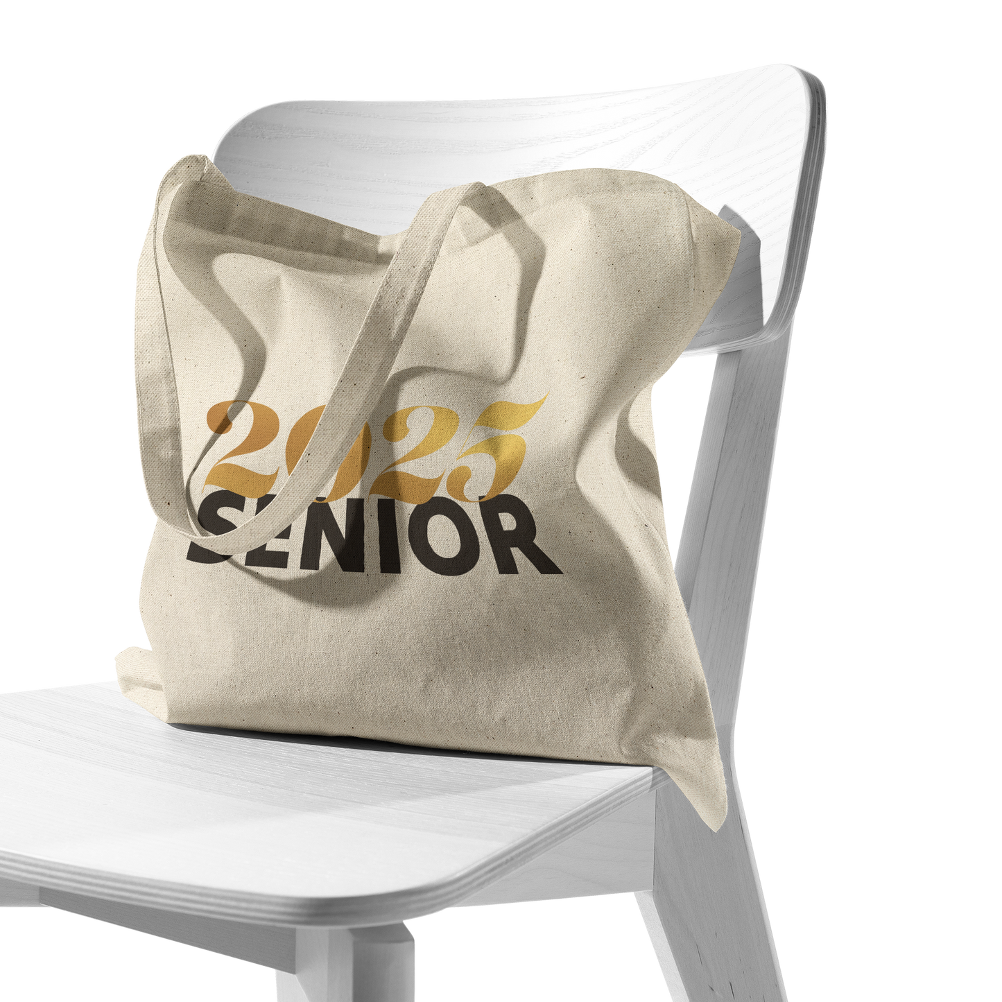 Tan canvas tote bag with 2025 in gold and senior in black sitting in a white chair.