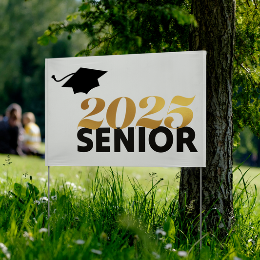 White vinyl yard sign with 2025 in gold, Senior in black text, and a black graduation cap in the top left placed in a park.