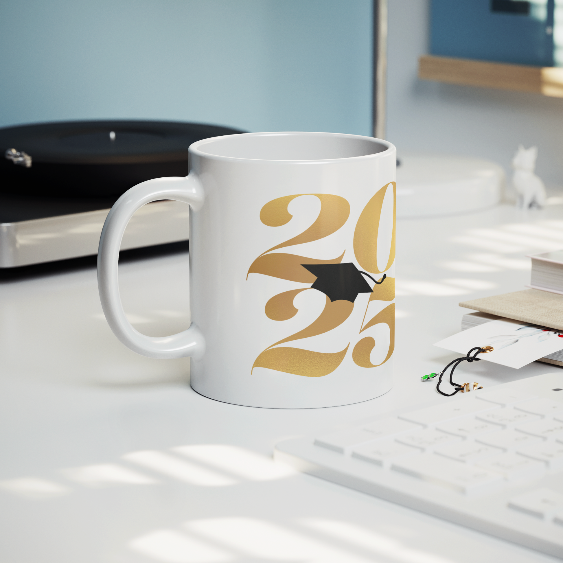 11oz white mug 2025 in gold text and a black graduation cap in the middle sitting on a desk with a keyboard, book, and record player.