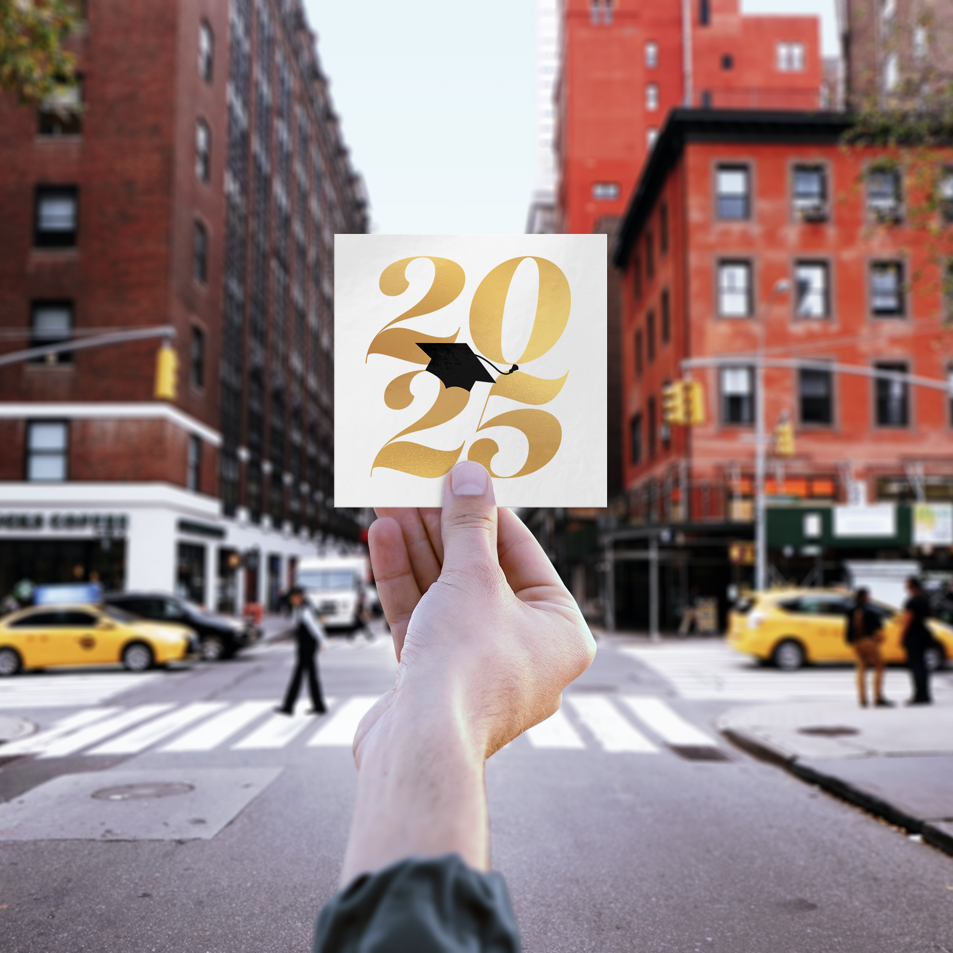 White square sticker with 2025 in gold and a black graduation cap placed in the middle being held up in front of a blurred city scape.