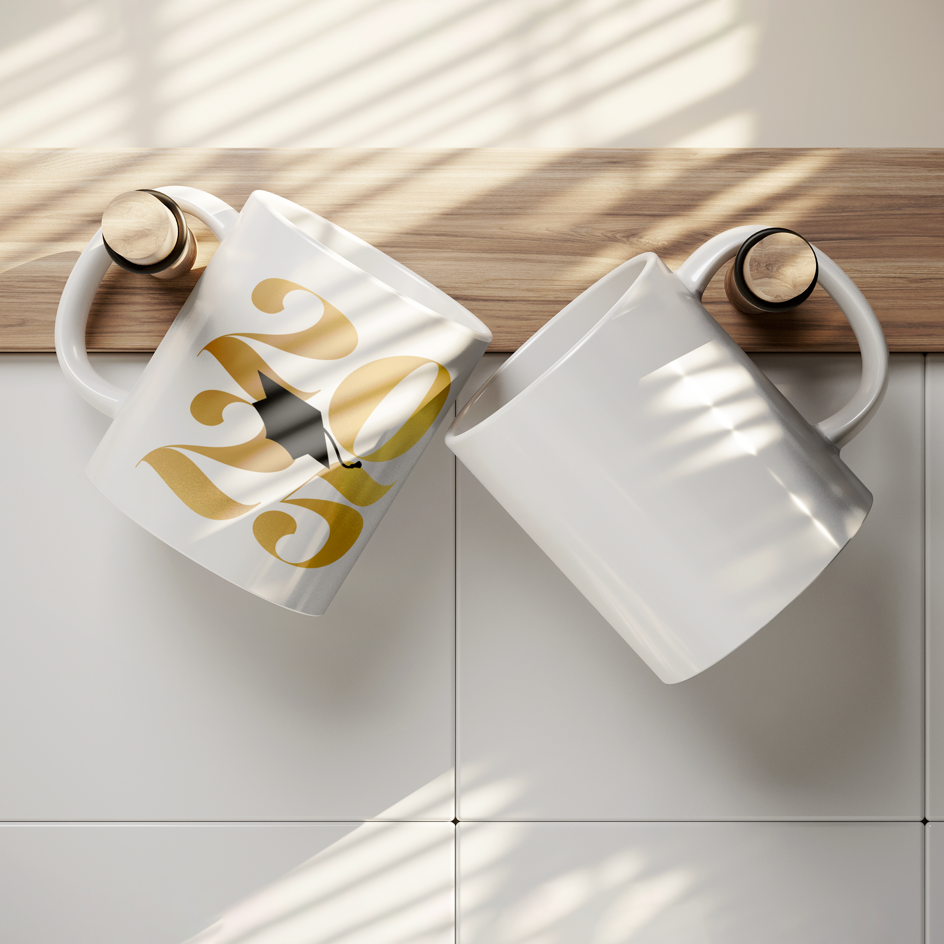 11oz white mug with 2025 in gold text and a black graduation cap in the middle hanging from a hook on a wall next to a blank white mug.