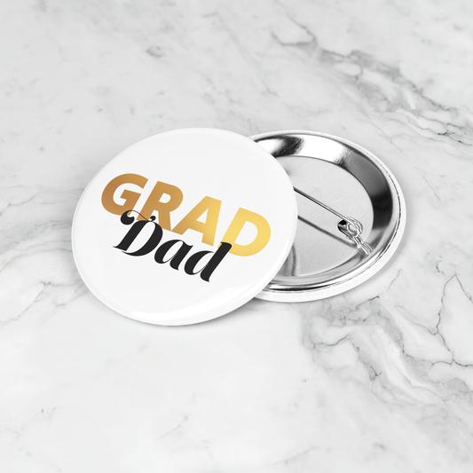 White button pin with Grad in gold and dad in black, laying on a marble surface.
