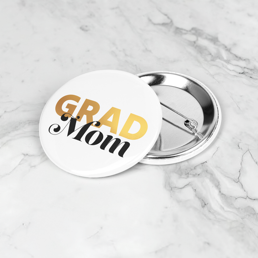 White button pin with Grad in gold and mom in black, laying on a marble surface.