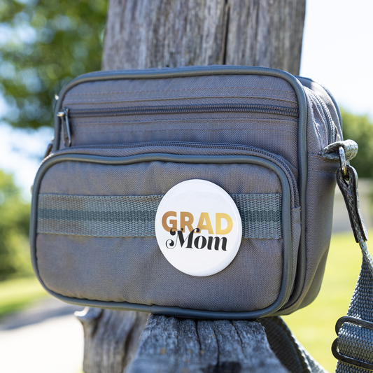 White button pin with Grad in gold and mom in black, pinned to a small gray bag sitting on a wooden bench. 