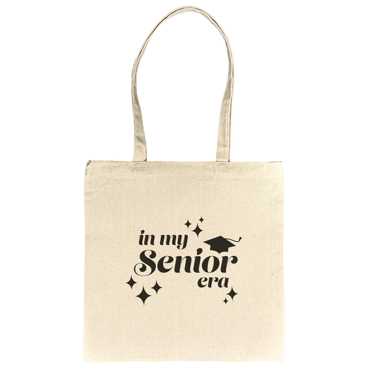 Tan canvas tote bag with the phrase “In My Senior Era” and a graduation cap printed in black ink laying flat