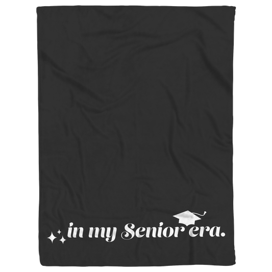 A black fleece blanket with the phrase "In My Senior Era" and a graduation cap in white at the bottom laying flat.