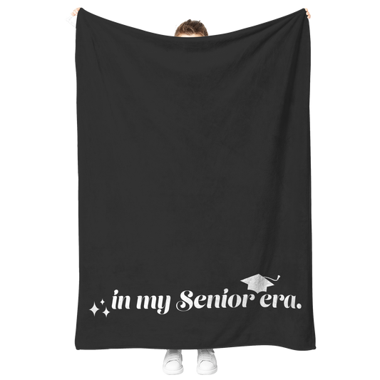 A black fleece blanket with the phrase "In My Senior Era" and a graduation cap in white at the bottom being held up.