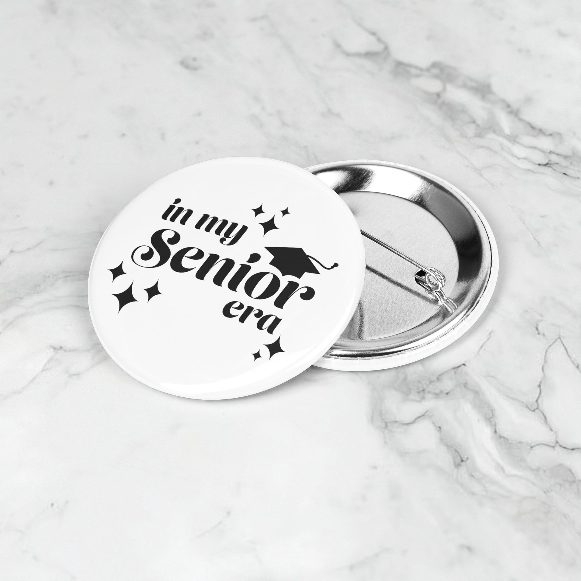 White button pin with the phrase "In My Senior Era" and a graduation cap in black sitting on a marble surface.