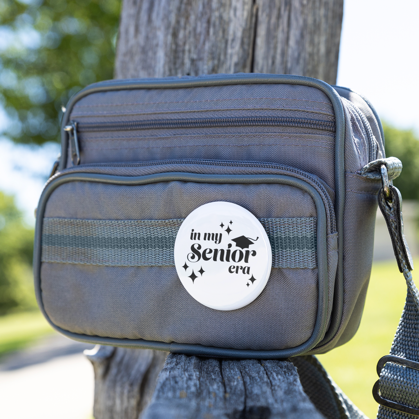 White button pin with the phrase "In My Senior Era" and a graduation cap in black pinned to a small gray bag sitting on a wooden bench. 