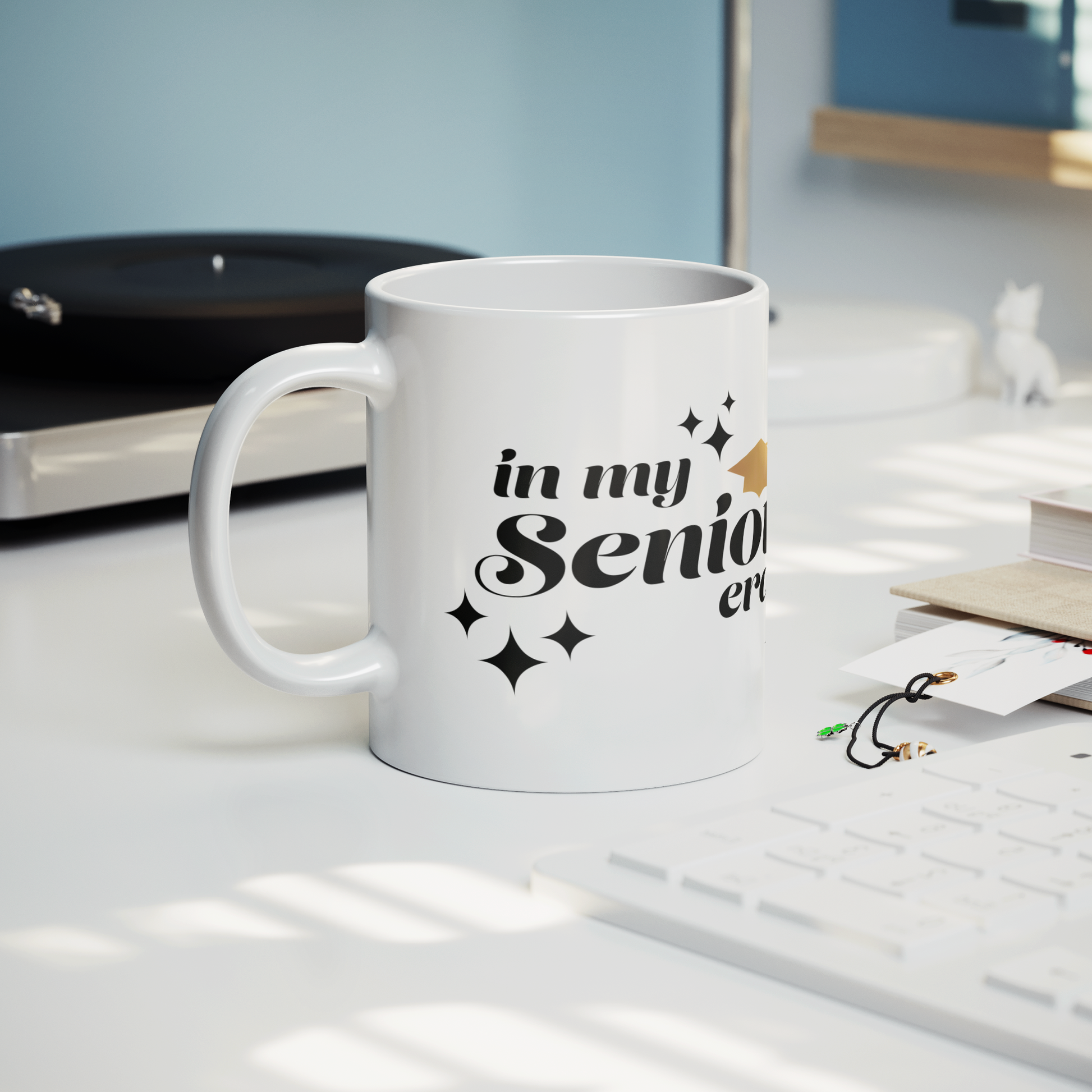 11oz white mug with the phrase “In My Senior Era” in black ink and a gold graduation cap sitting on a desk with a keyboard, book, and record player.