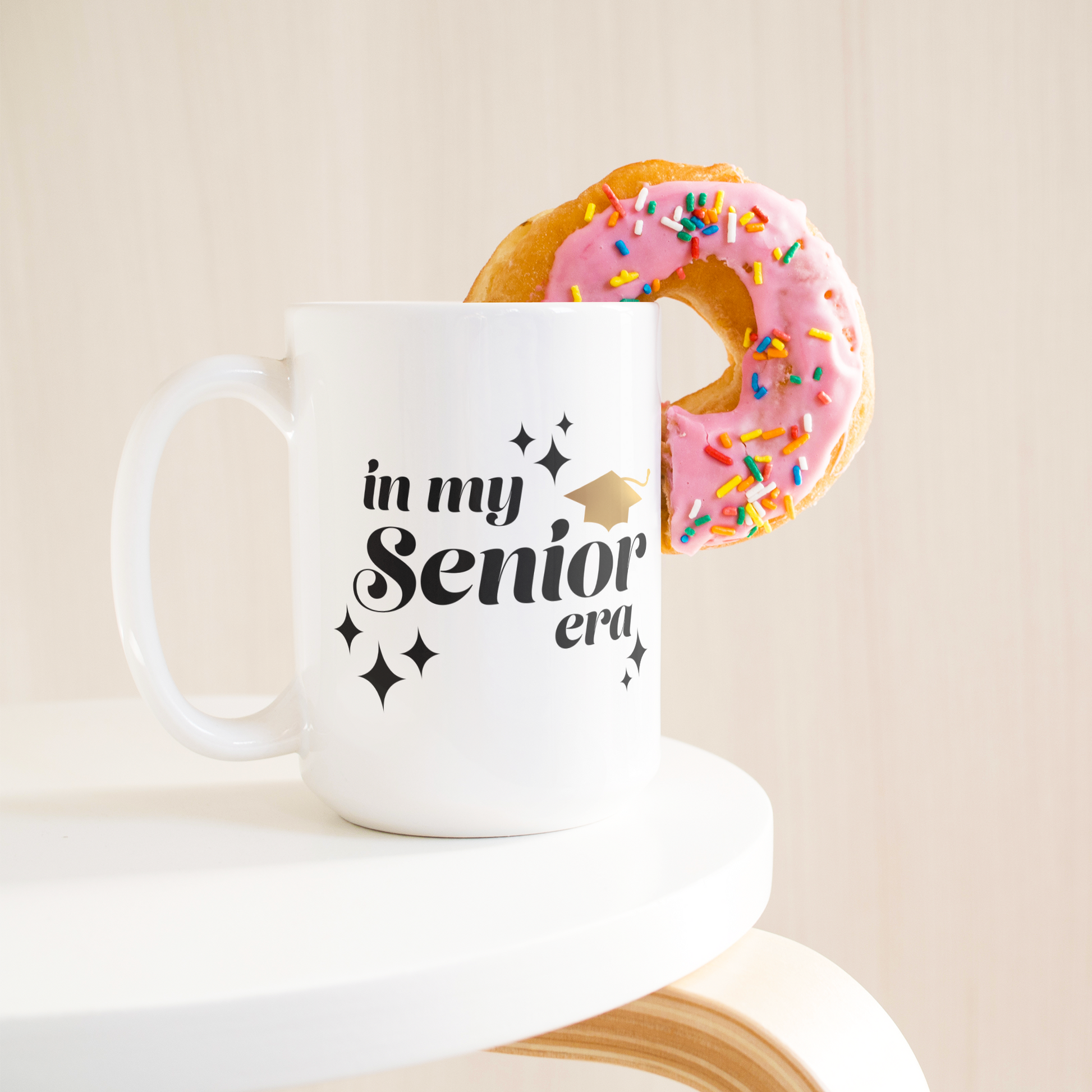 15oz white mug with the text "In My Senior Era" in black and a gold graduation cap sitting on a table with a donut sticking out of the mug.