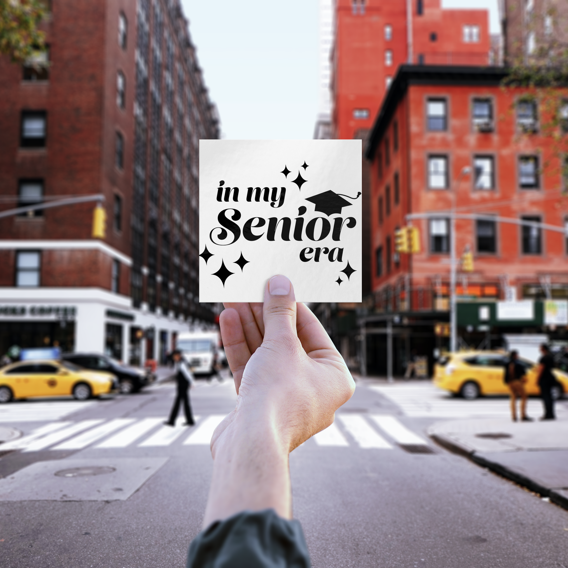 White square sticker with the phrase “In My Senior Era” and a graduation cap in black ink being held up in front of a blurred city scape.