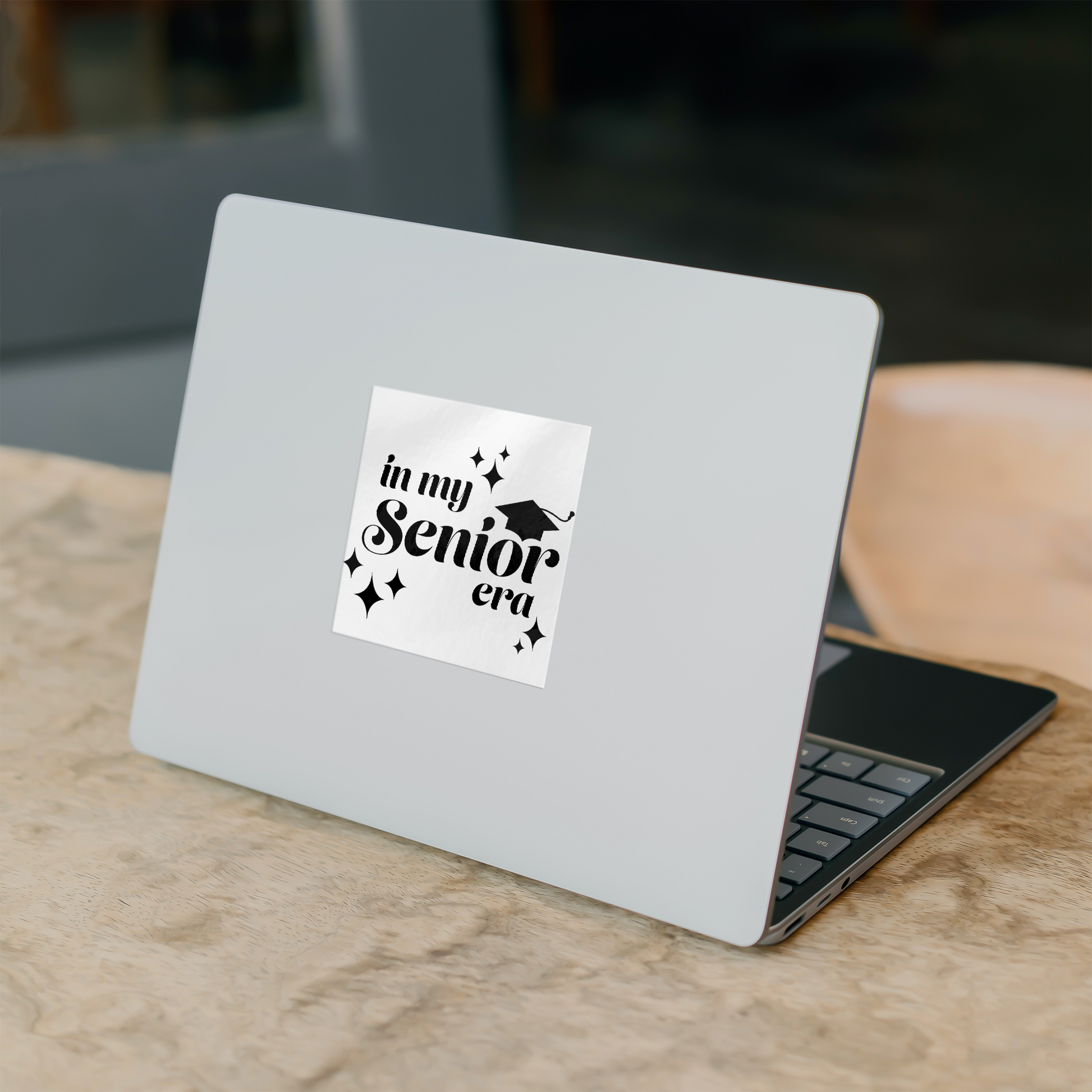 White square sticker with the phrase “In My Senior Era” and a graduation cap in black ink stuck in the middle of a silver laptop on a desk.