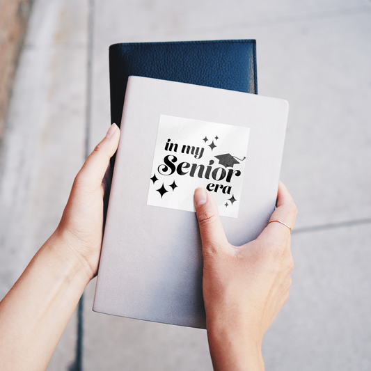 White square sticker with the phrase “In My Senior Era” and a graduation cap in black ink stuck on a gray notebook being held.