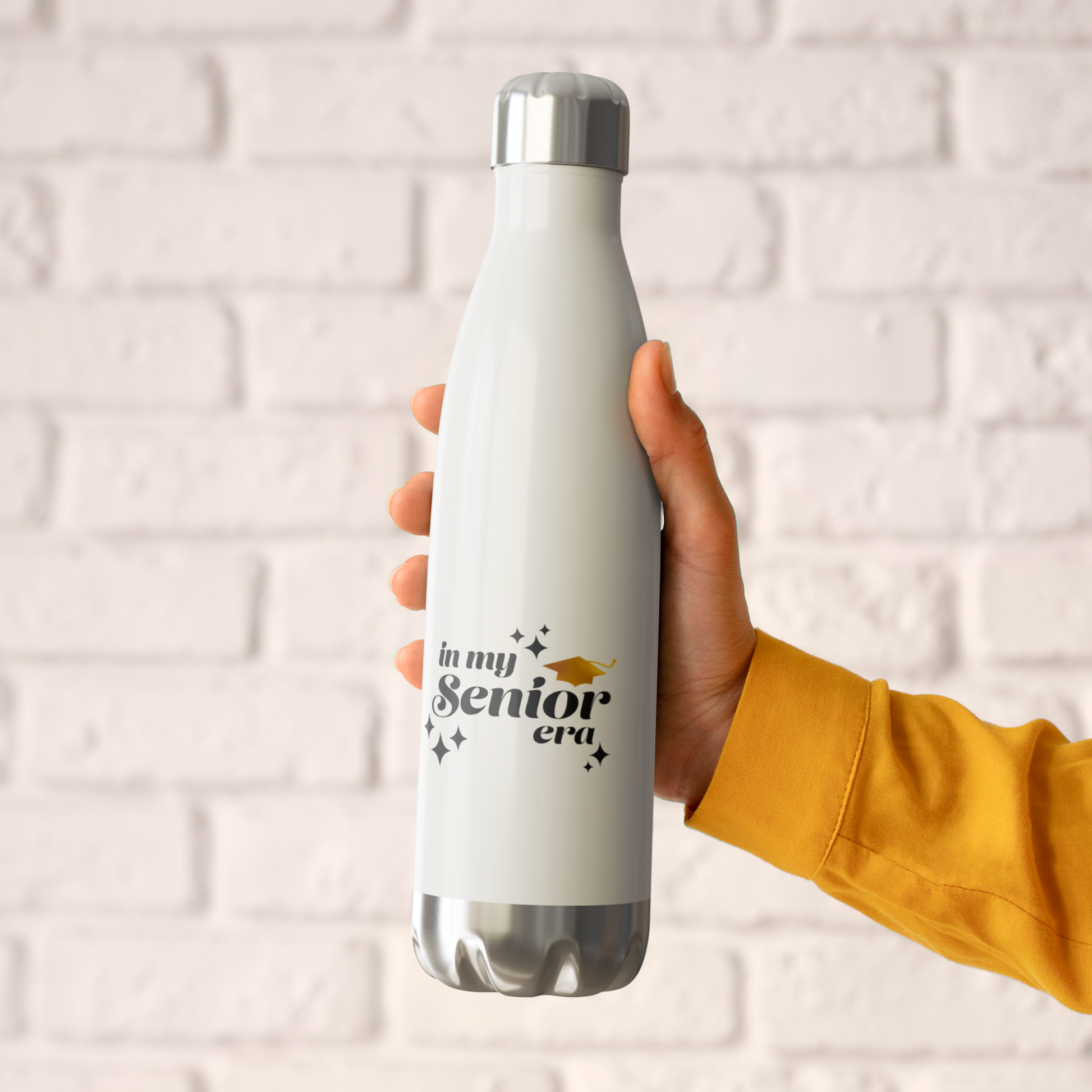 A white, 20oz water bottle with a silver lid and base being held by a hand in front of a white brick wall. Text in the bottom center of the bottle says "In my Senior Era" in black with a gold graduation cap.